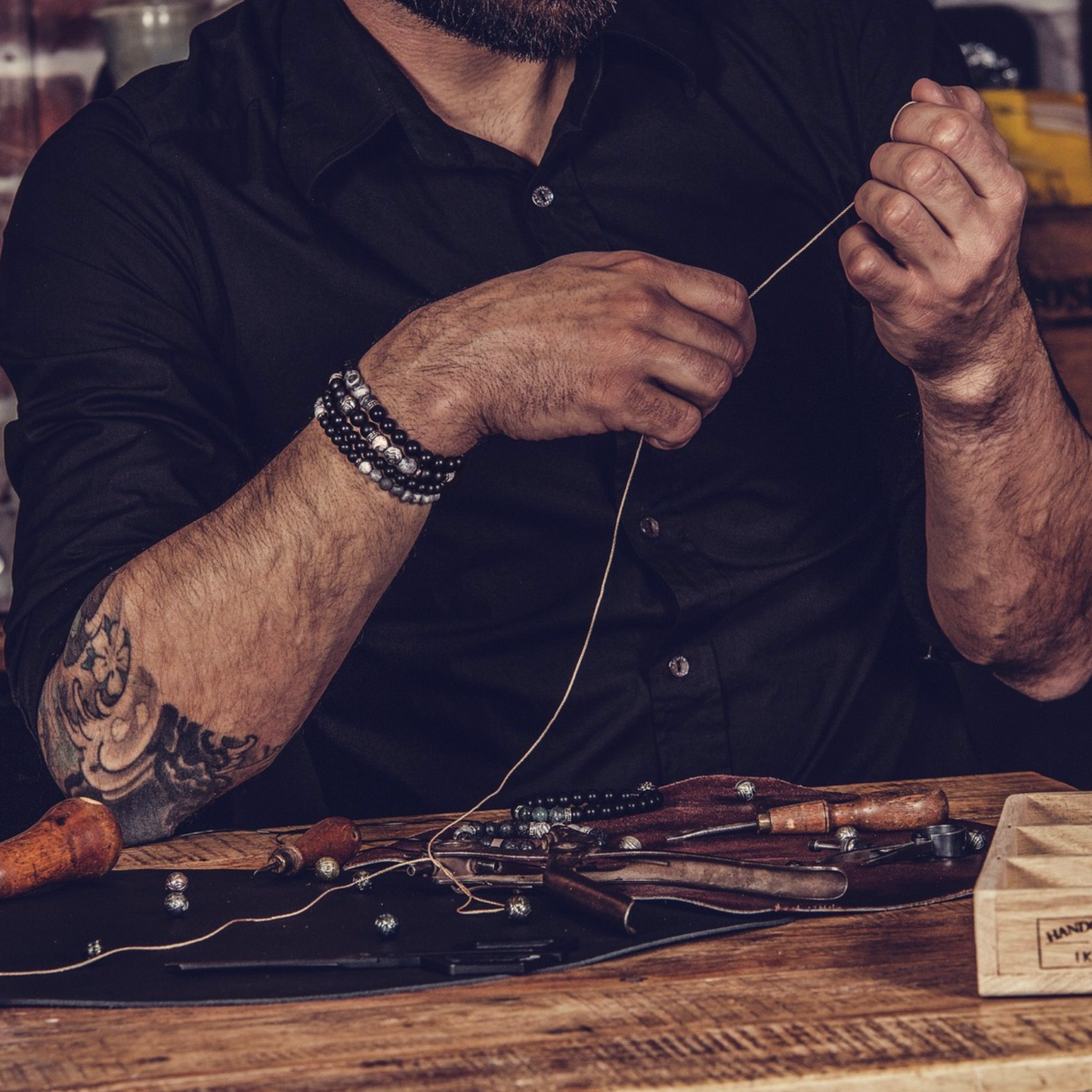 A close-up lifestyle shot of a bearded man wearing a black shirt, carefully threading a needle with both hands. He is wearing a stack of black and silver beaded bracelets on his left wrist.