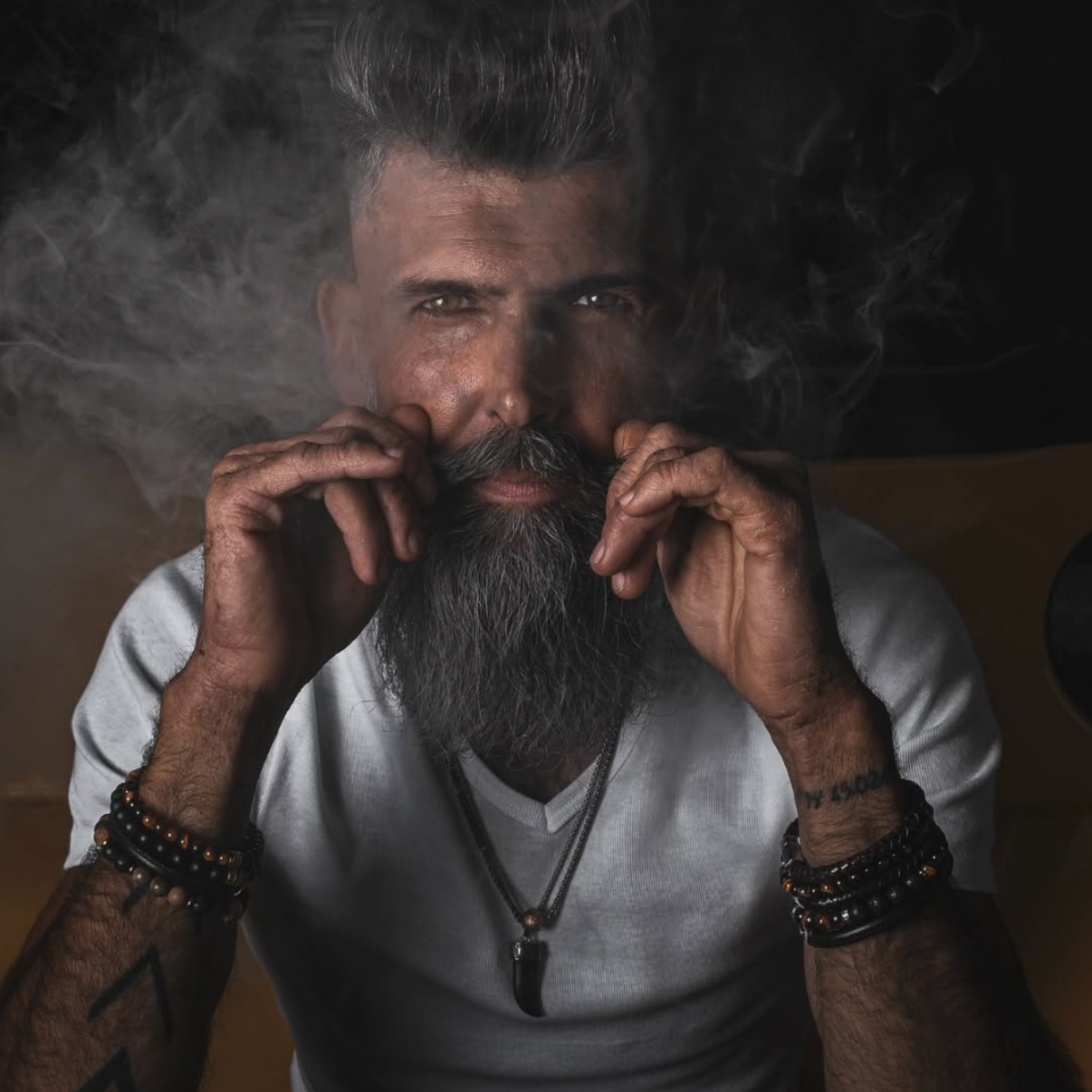 A close-up portrait of a man with a long gray beard and styled hair, wearing a white t-shirt and black beaded bracelets, with smoke swirling around his face against a dark background.