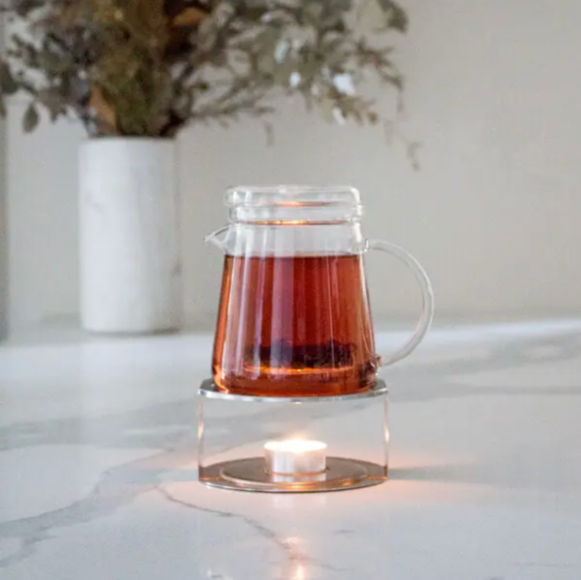 Clear glass T42 teapot with tea on a marble surface with a candle and plant in the background